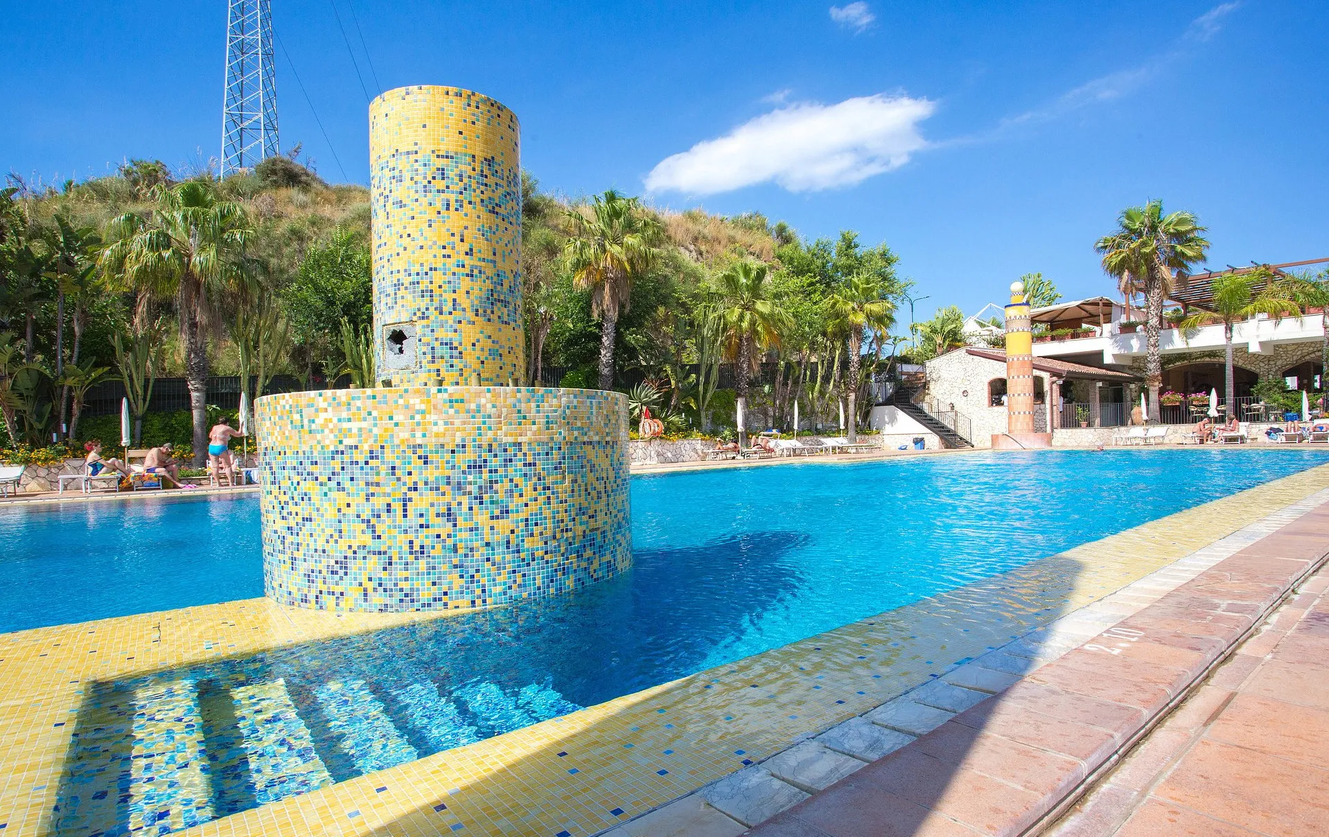 Dettaglio della piscina con colonne a mosaico gialle e blu e palme
