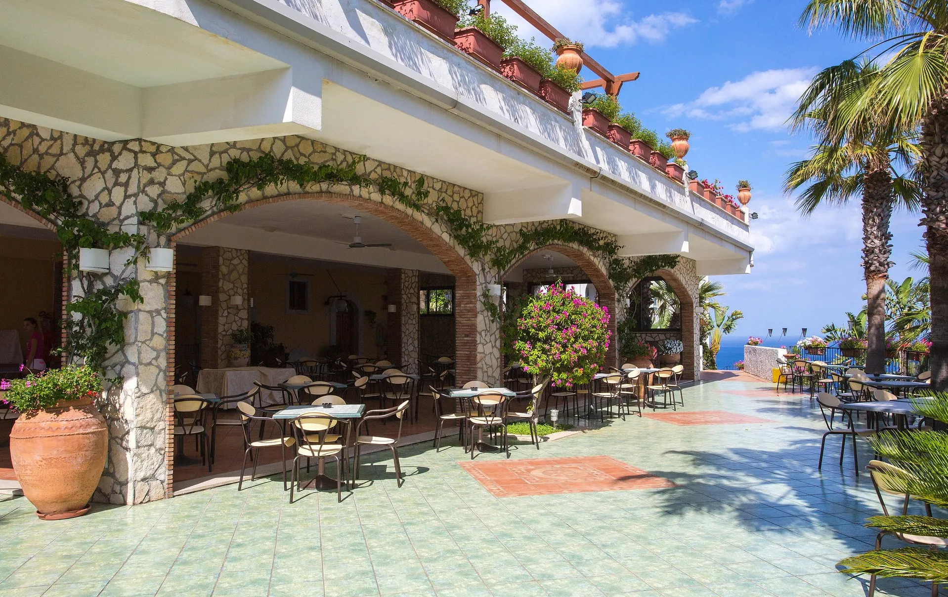 Terrazza coperta con archi in pietra, tavoli e vista sul mare tra le palme