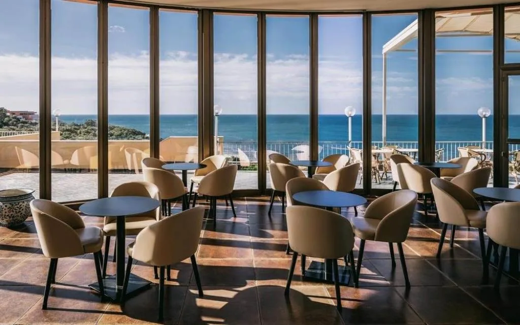 Sala lounge interna con poltrone, tavolini e grandi vetrate sul mare