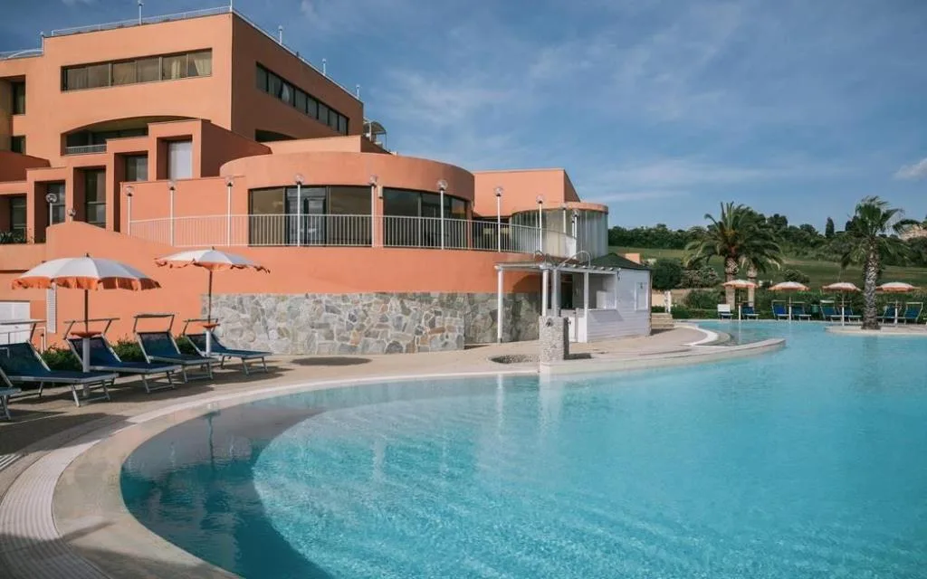 Piscina esterna curva con lettini, ombrelloni e struttura dell'hotel color corallo