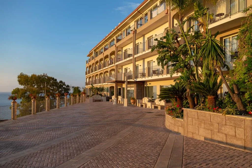 Facciata dell'hotel con terrazza pavimentata e vegetazione mediterranea al tramonto
