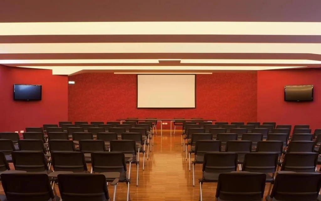 Sala conferenze con pareti rosse, sedie nere allineate, schermo per proiezioni e monitor
