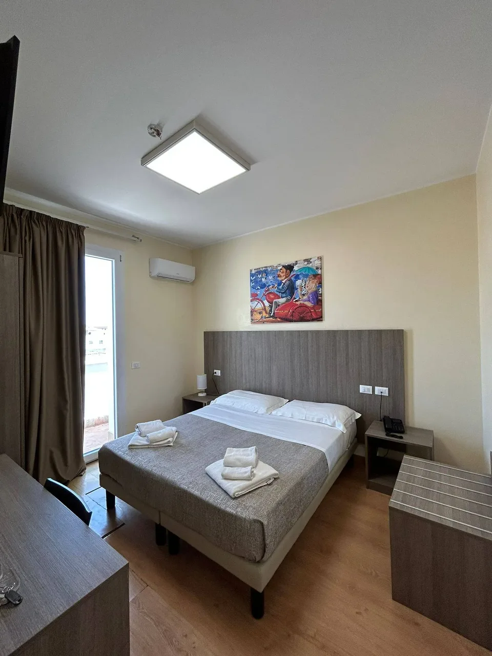 Camera matrimoniale luminosa con porta finestra, scrivania e quadro decorativo