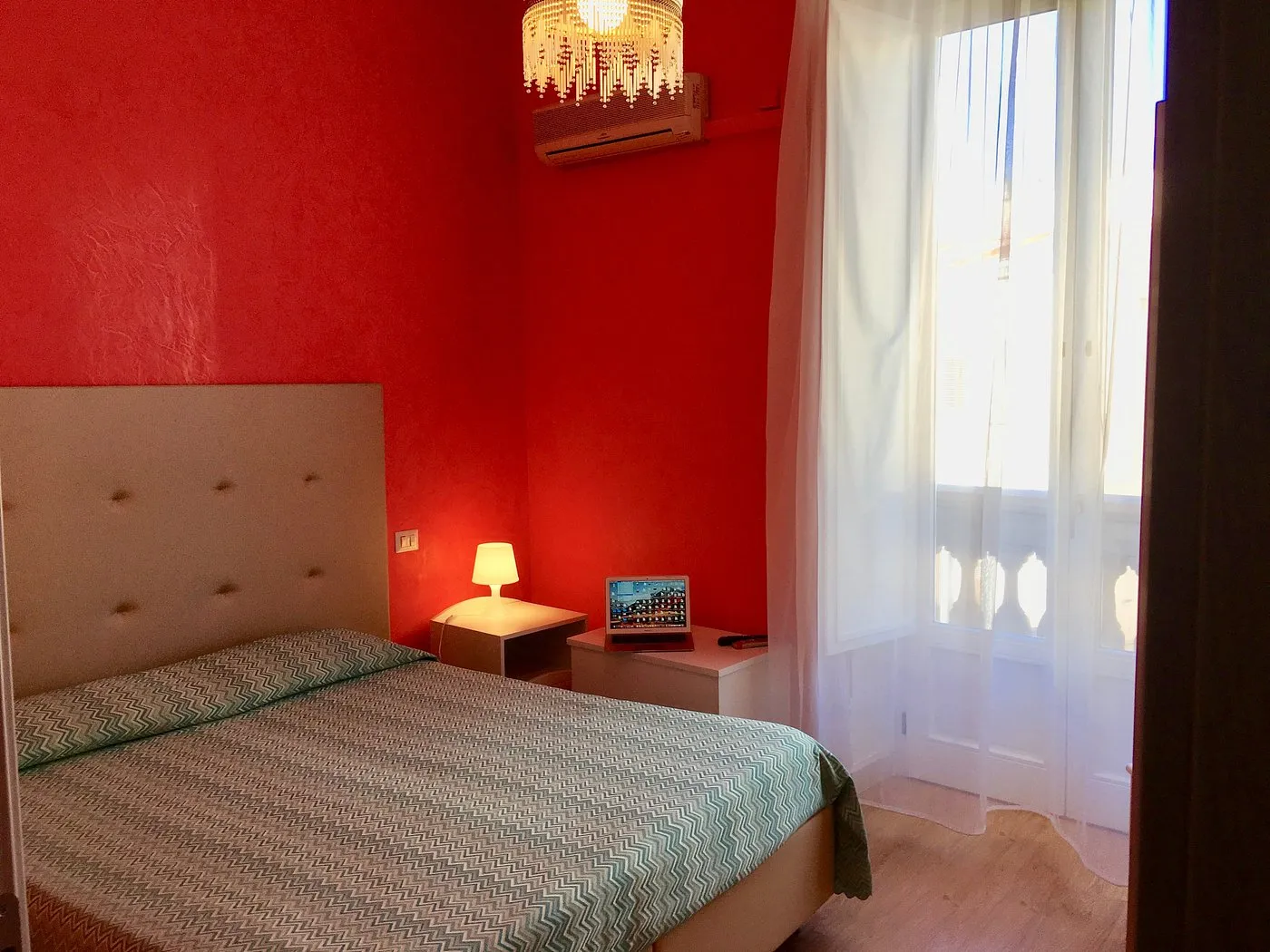 Camera con parete rossa, letto matrimoniale, finestra luminosa con tende bianche e aria condizionata