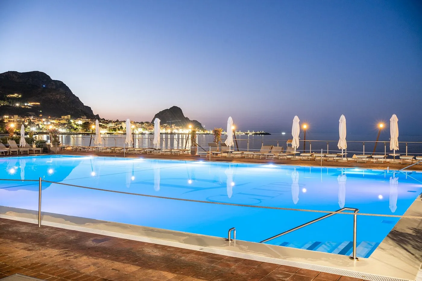 Piscina serale affacciata sul mare con vista sulle luci della città e le montagne