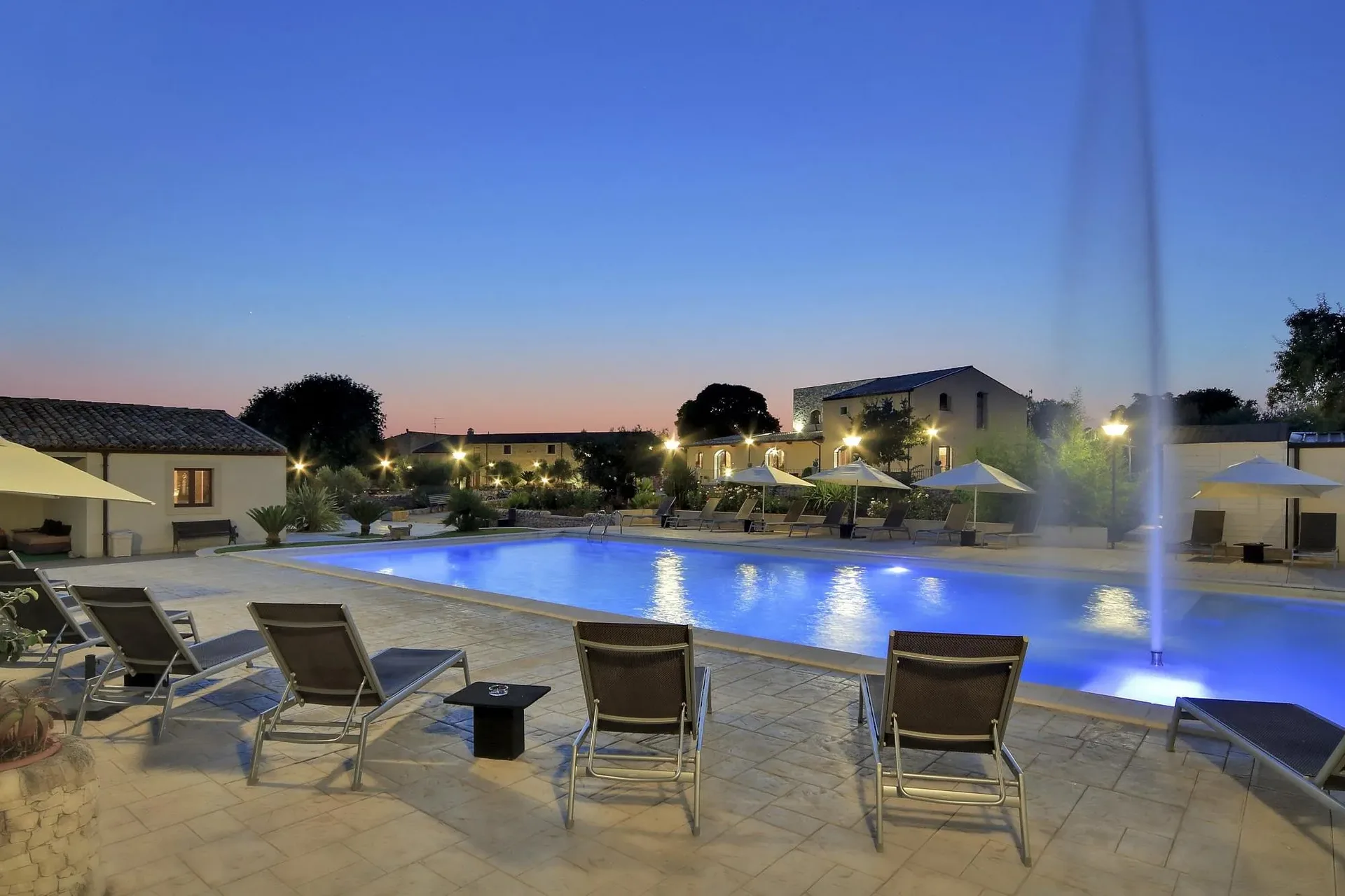 Piscina illuminata al crepuscolo con lettini, ombrelloni e getto d'acqua