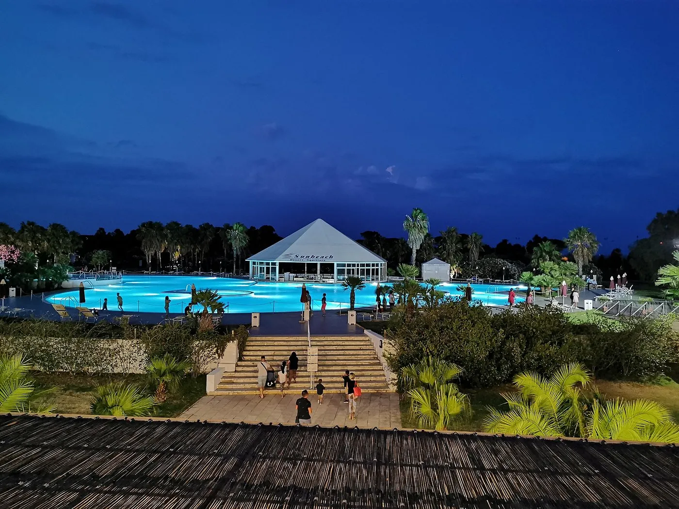 Piscina illuminata di sera con gazebo bianco e persone che scendono le scale