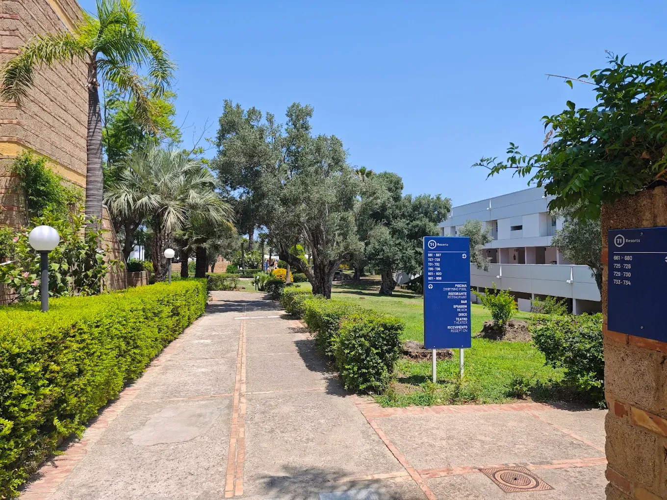 Vialetto del resort con giardino curato, palme, ulivi e cartelli indicativi blu