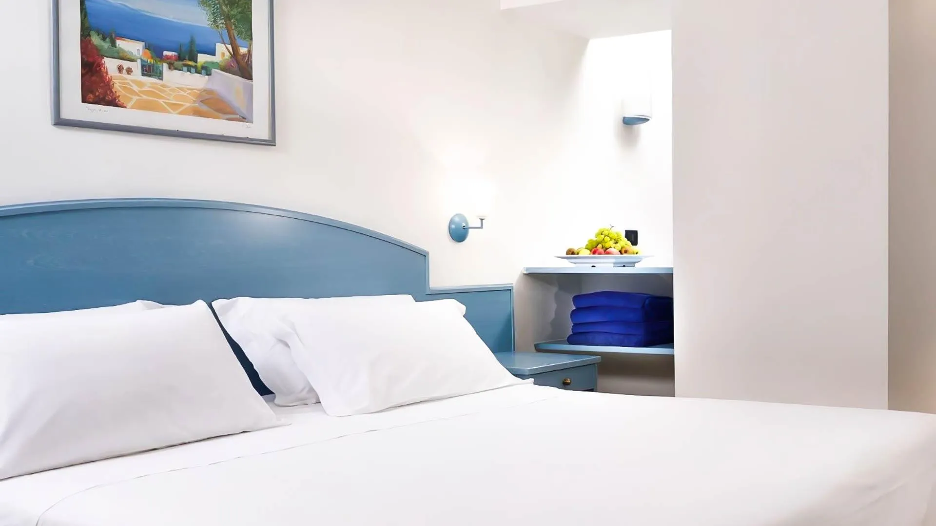 Dettaglio di camera con testiera azzurra, letto bianco e comodino con frutta fresca