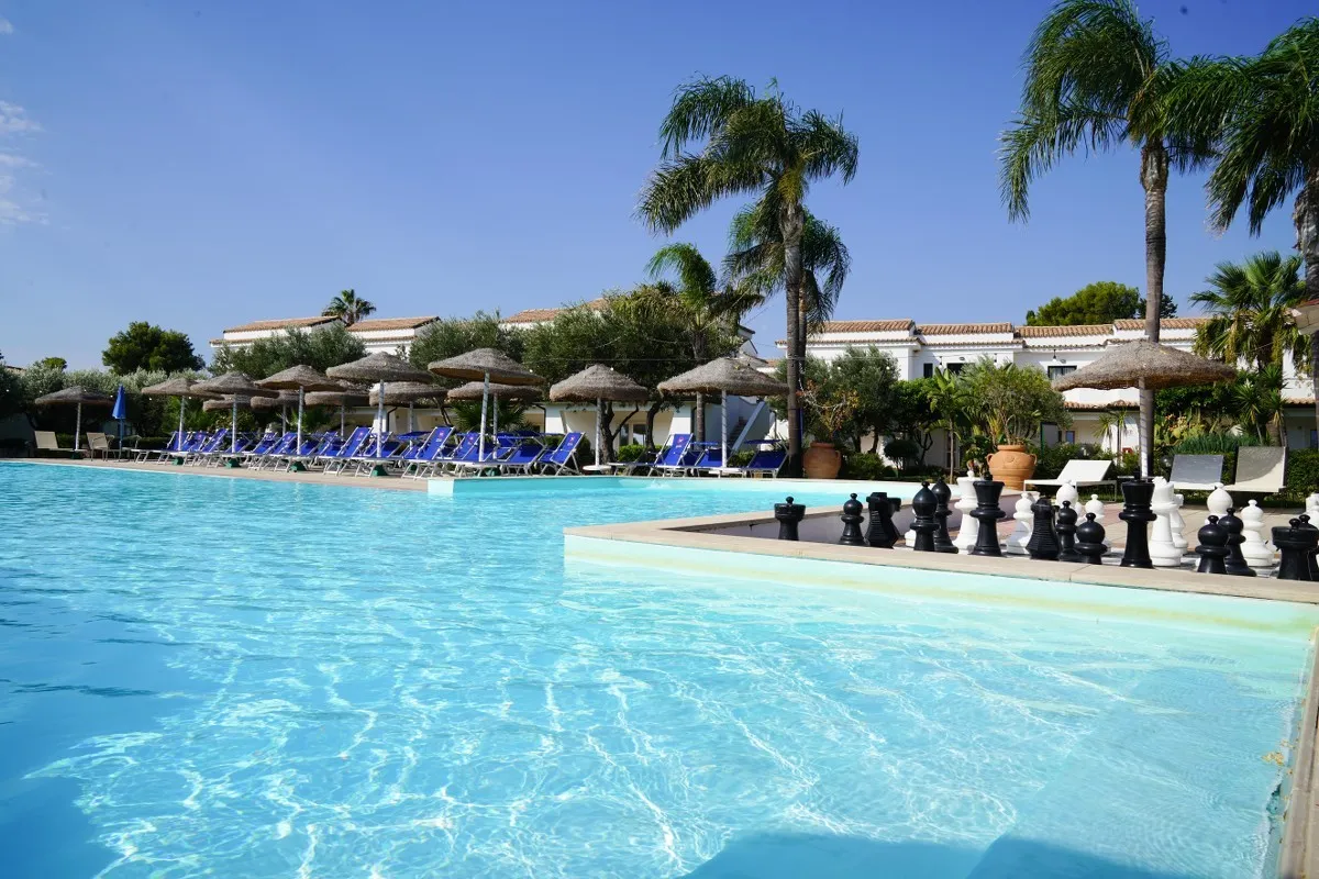 Piscina con lettini blu, ombrelloni in paglia e scacchiera gigante