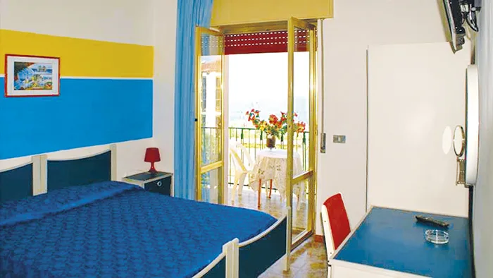 Camera matrimoniale con pareti gialle e blu, balcone con vista mare e tavolino fiorito