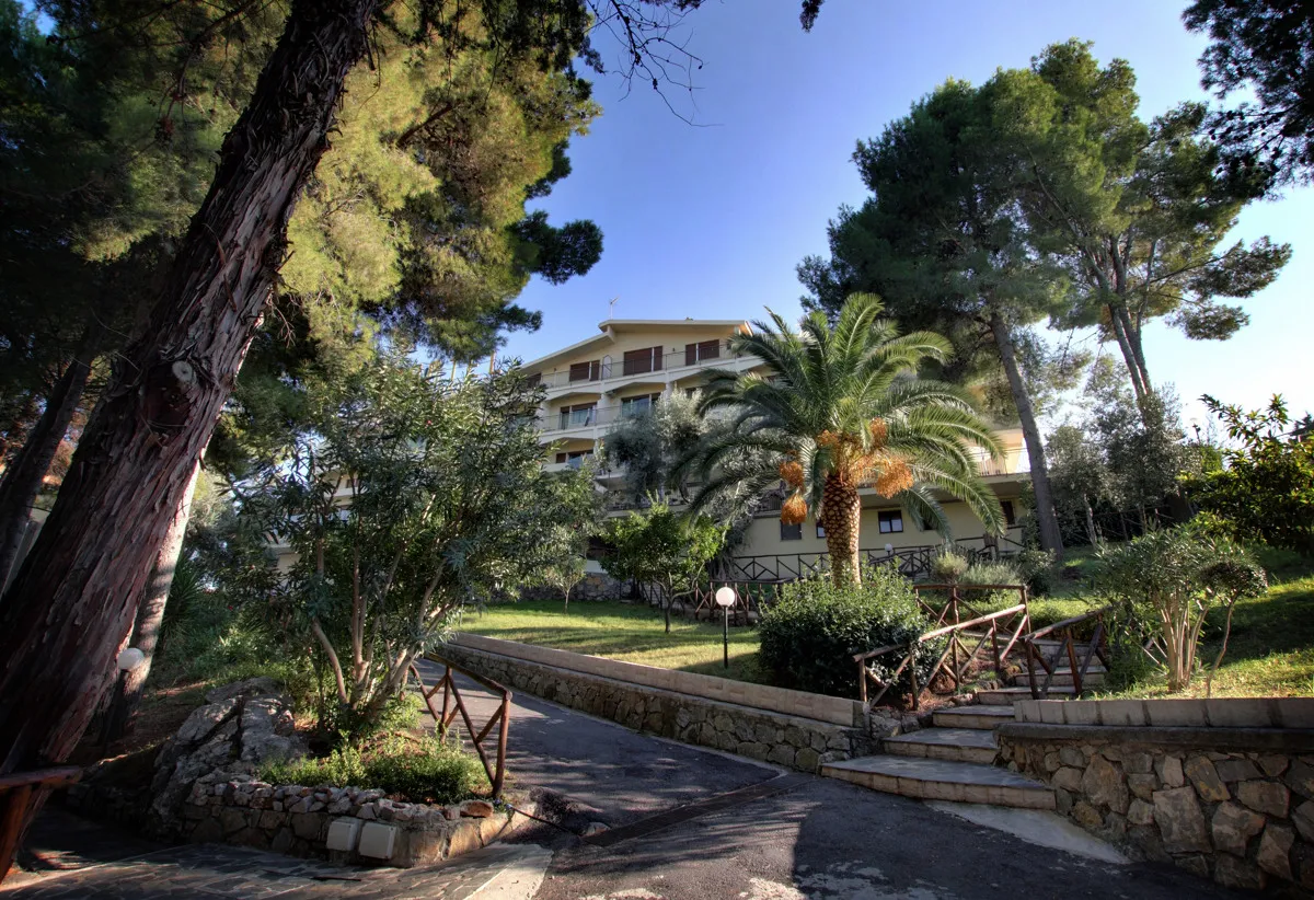 Edificio dell'hotel immerso nel verde tra pini e palme con vialetto e giardino