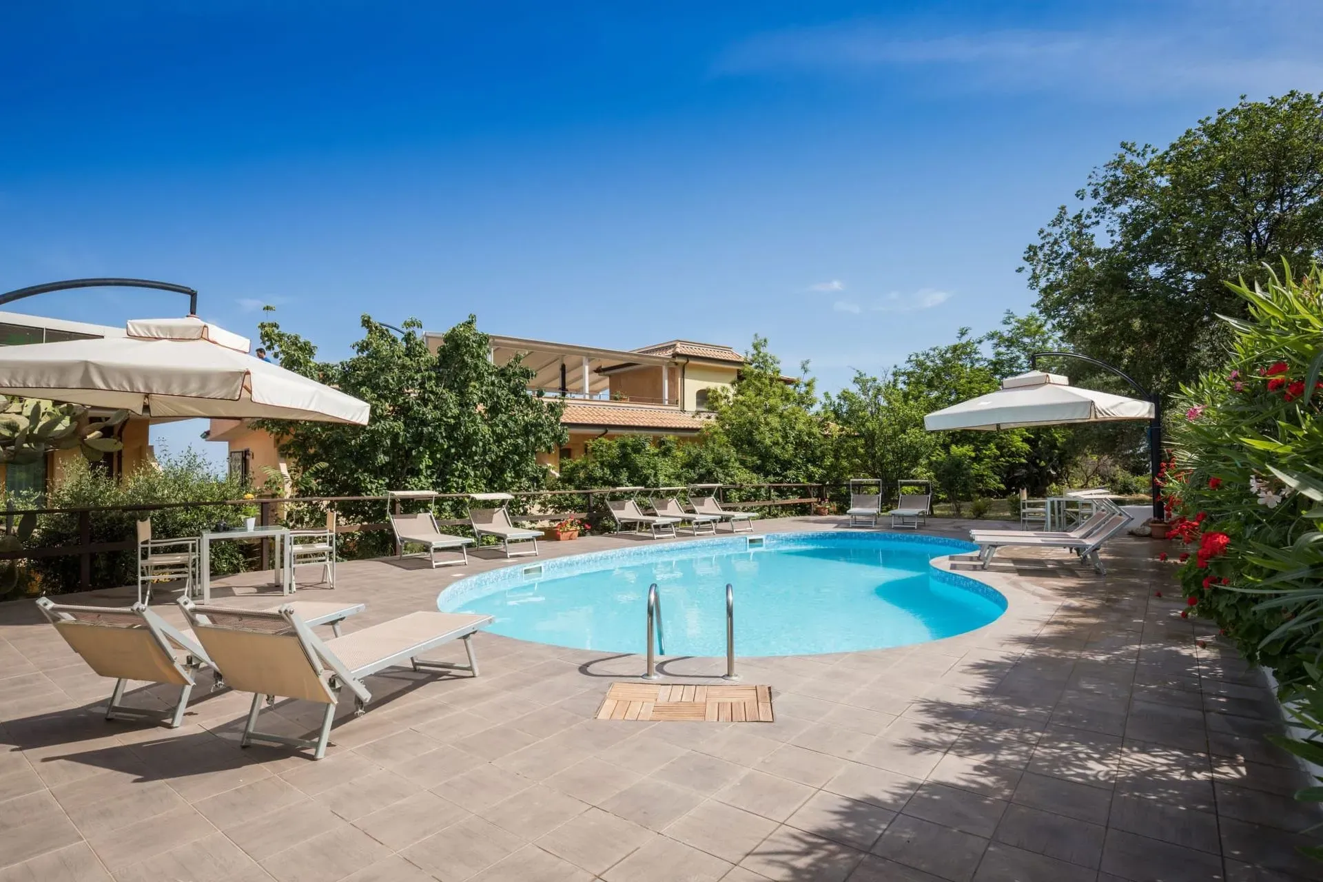 Piscina esterna con lettini, ombrelloni e giardino sotto il cielo azzurro