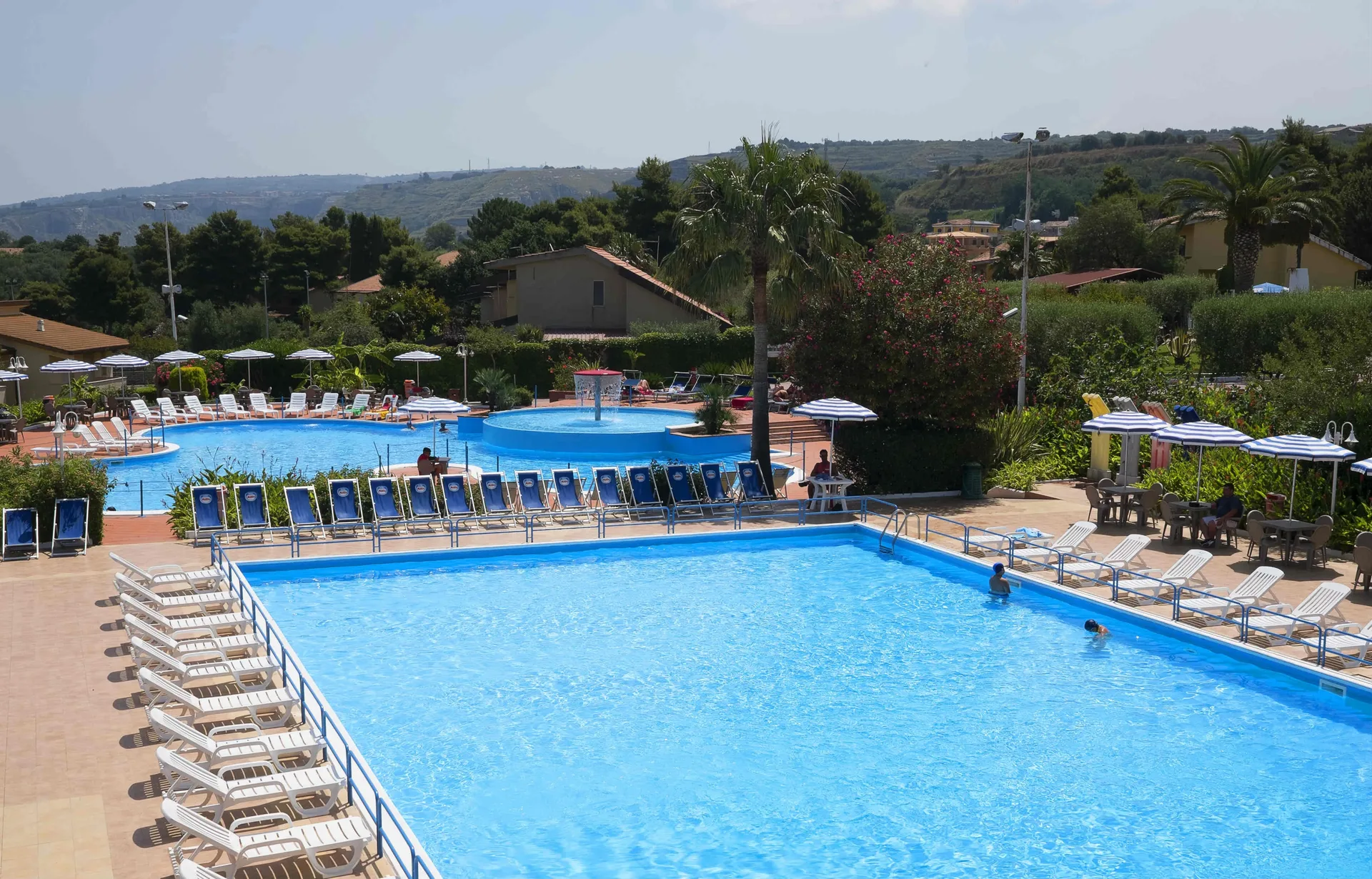 Piscine all'aperto con lettini, ombrelloni e palme con vista sulle colline