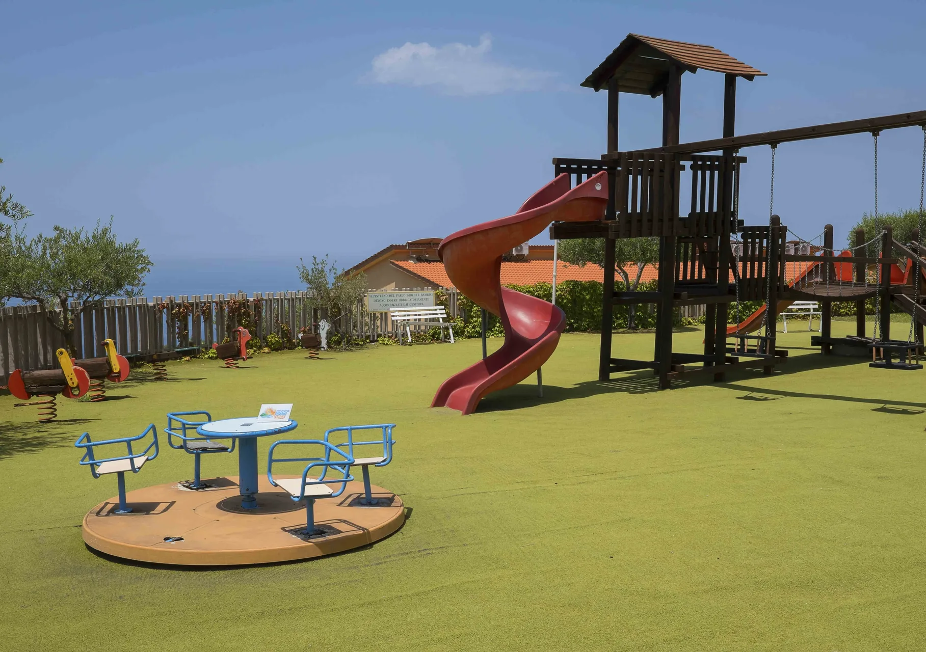 Area giochi per bambini con scivolo, altalena e giostrina su prato verde con vista mare