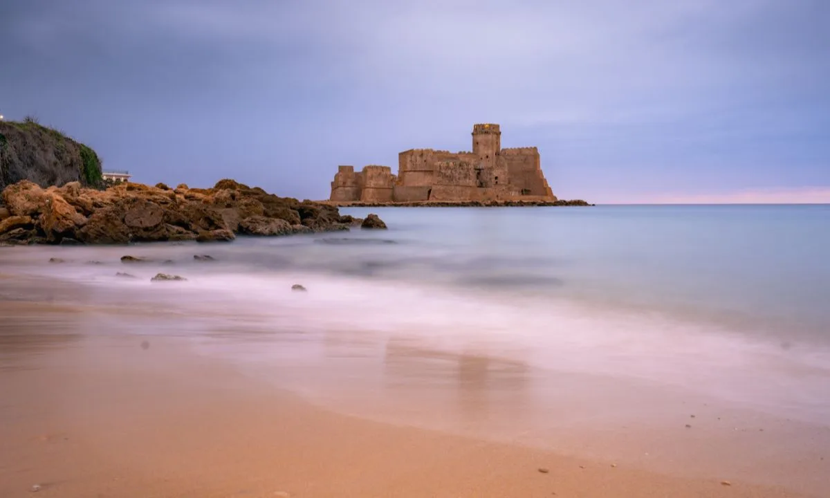 Spiaggia sabbiosa con scogli e antica fortezza sul mare all'orizzonte in atmosfera crepuscolare