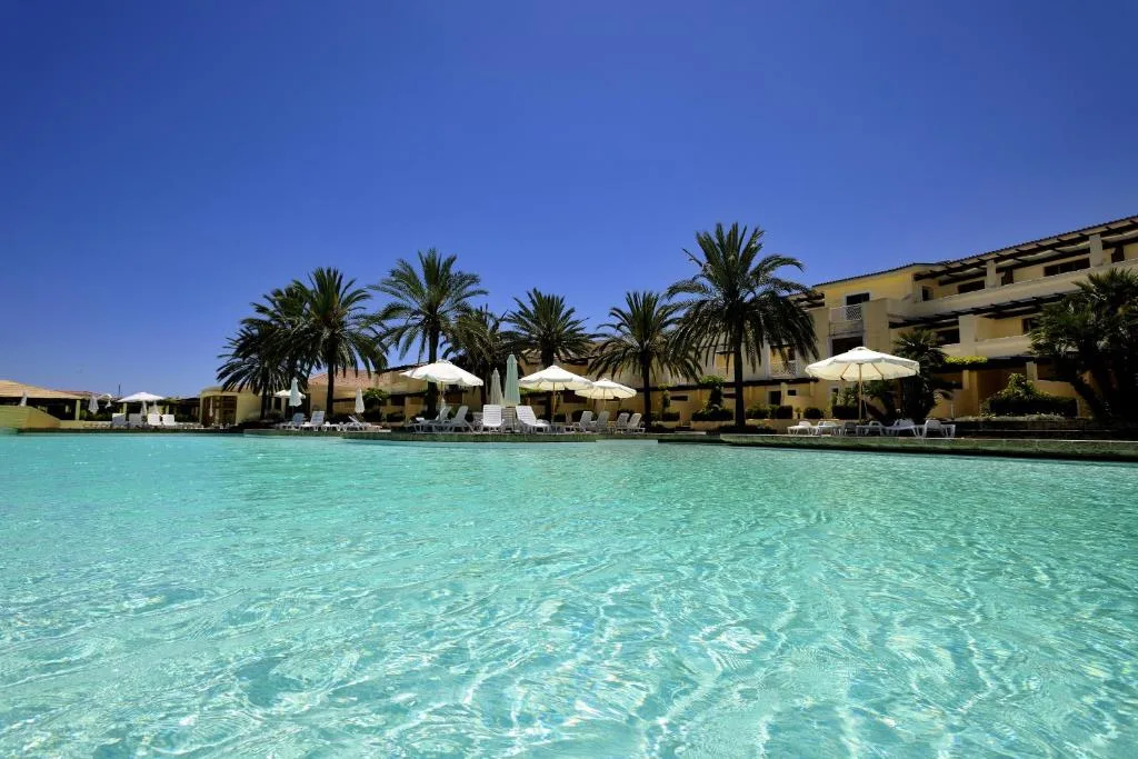 Piscina dalle acque trasparenti con palme, ombrelloni bianchi ed edifici del resort
