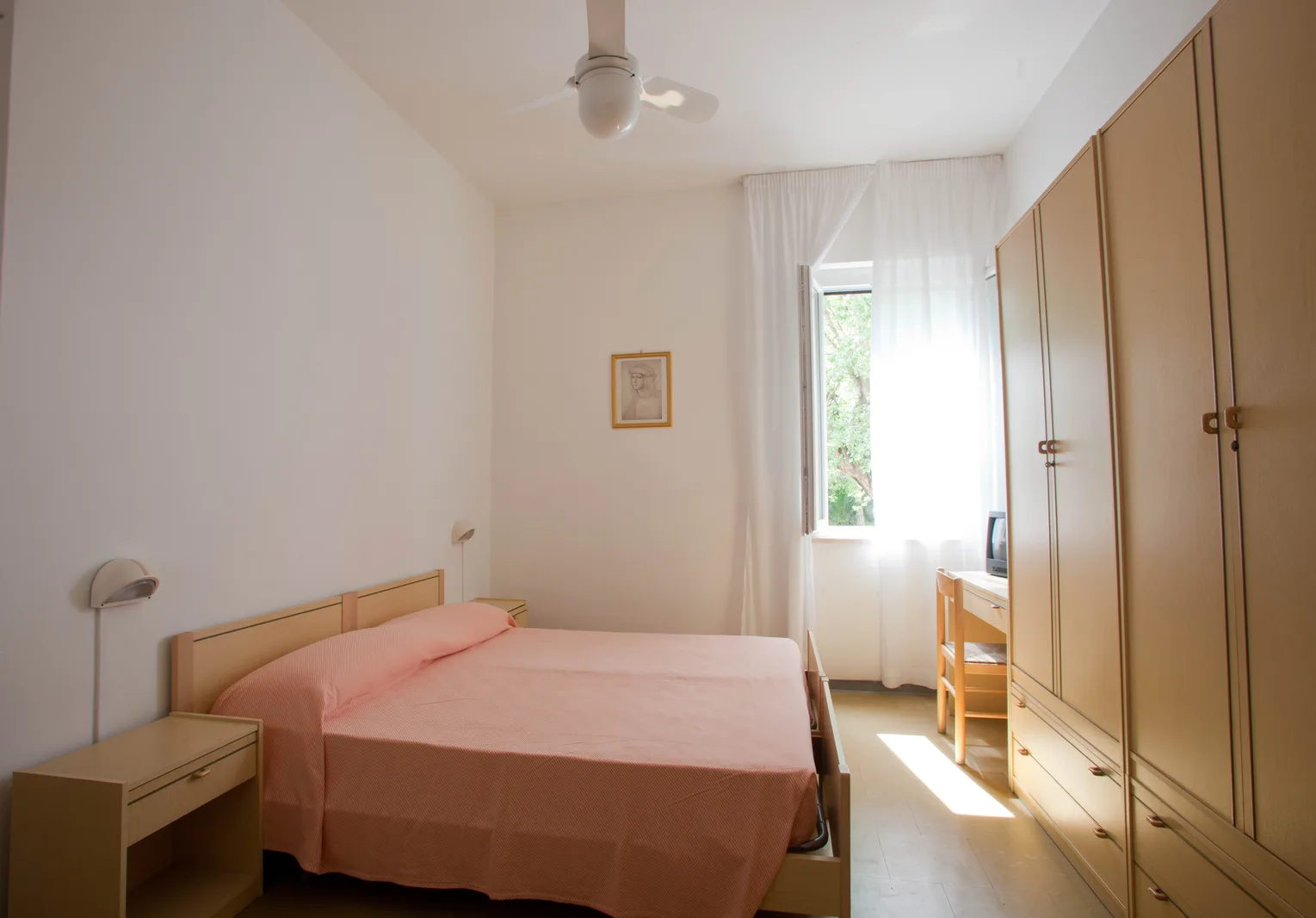 Camera da letto luminosa con copriletto rosa, armadio in legno chiaro, scrivania e finestra con vista sul verde