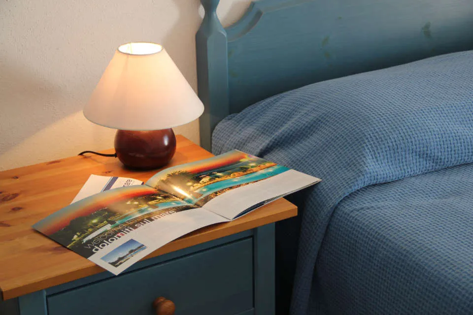 Dettaglio comodino in legno con lampada e brochure accanto al letto blu