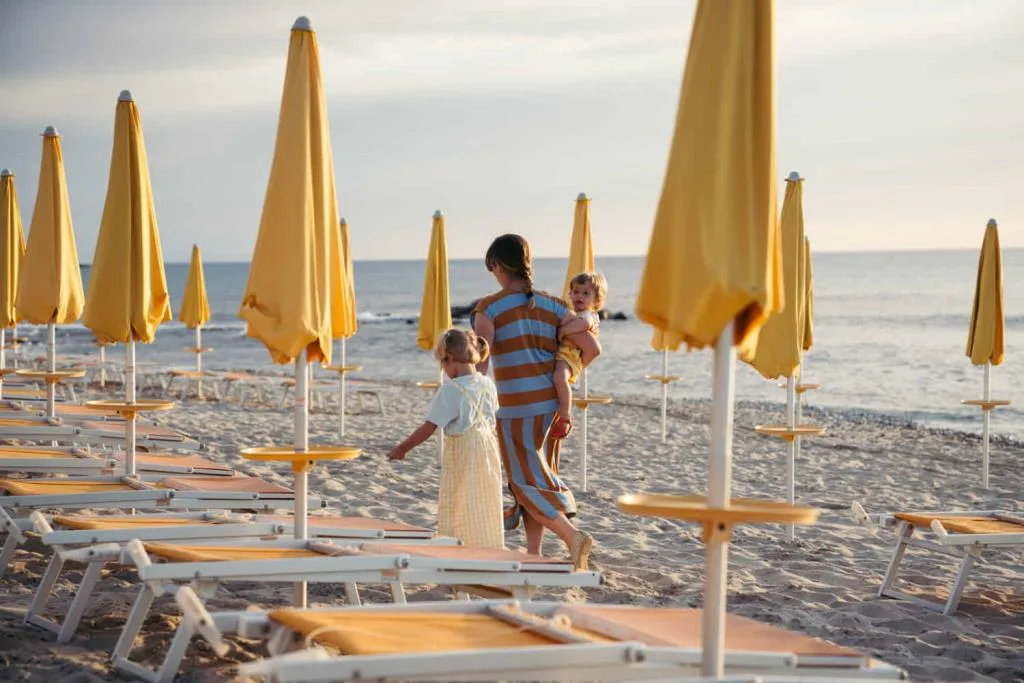 Mamma con due bambini cammina tra lettini e ombrelloni gialli chiusi sulla spiaggia al tramonto