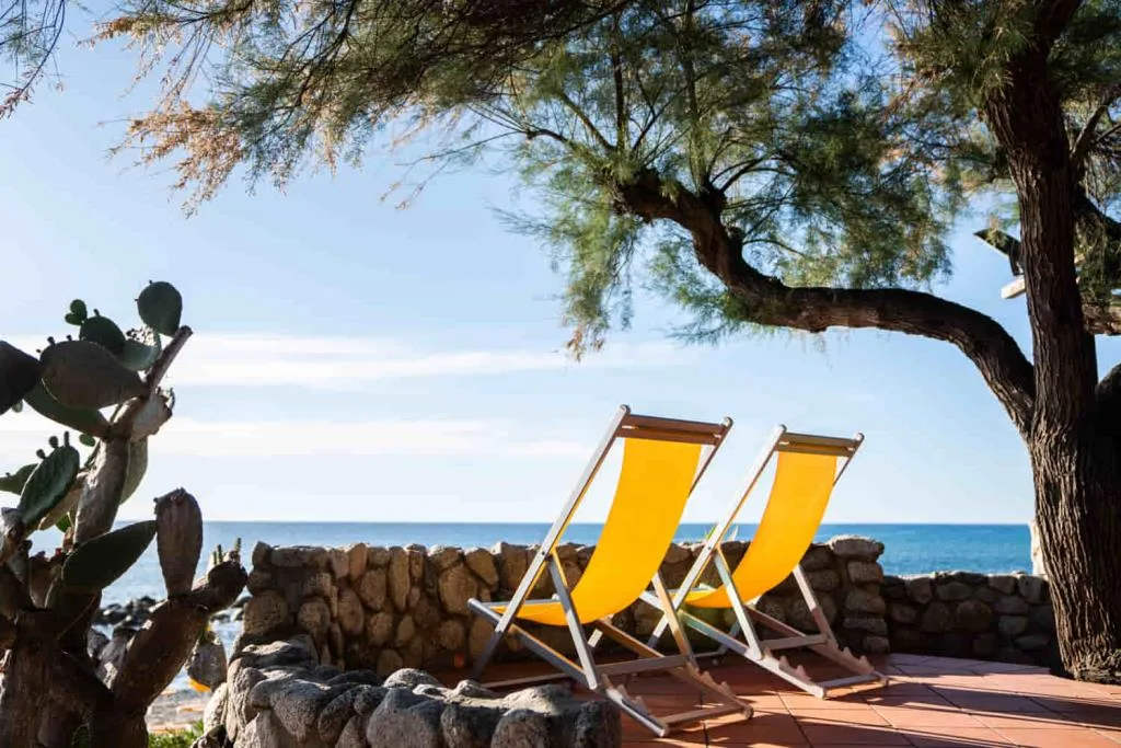 Due sdraio gialle all'ombra di un albero con vista sul mare e muretto in pietra