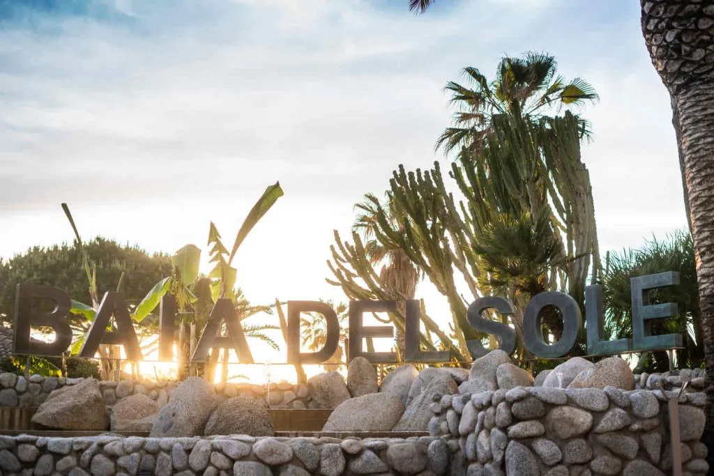 Insegna Baia del Sole in metallo tra rocce, cactus e palme al tramonto