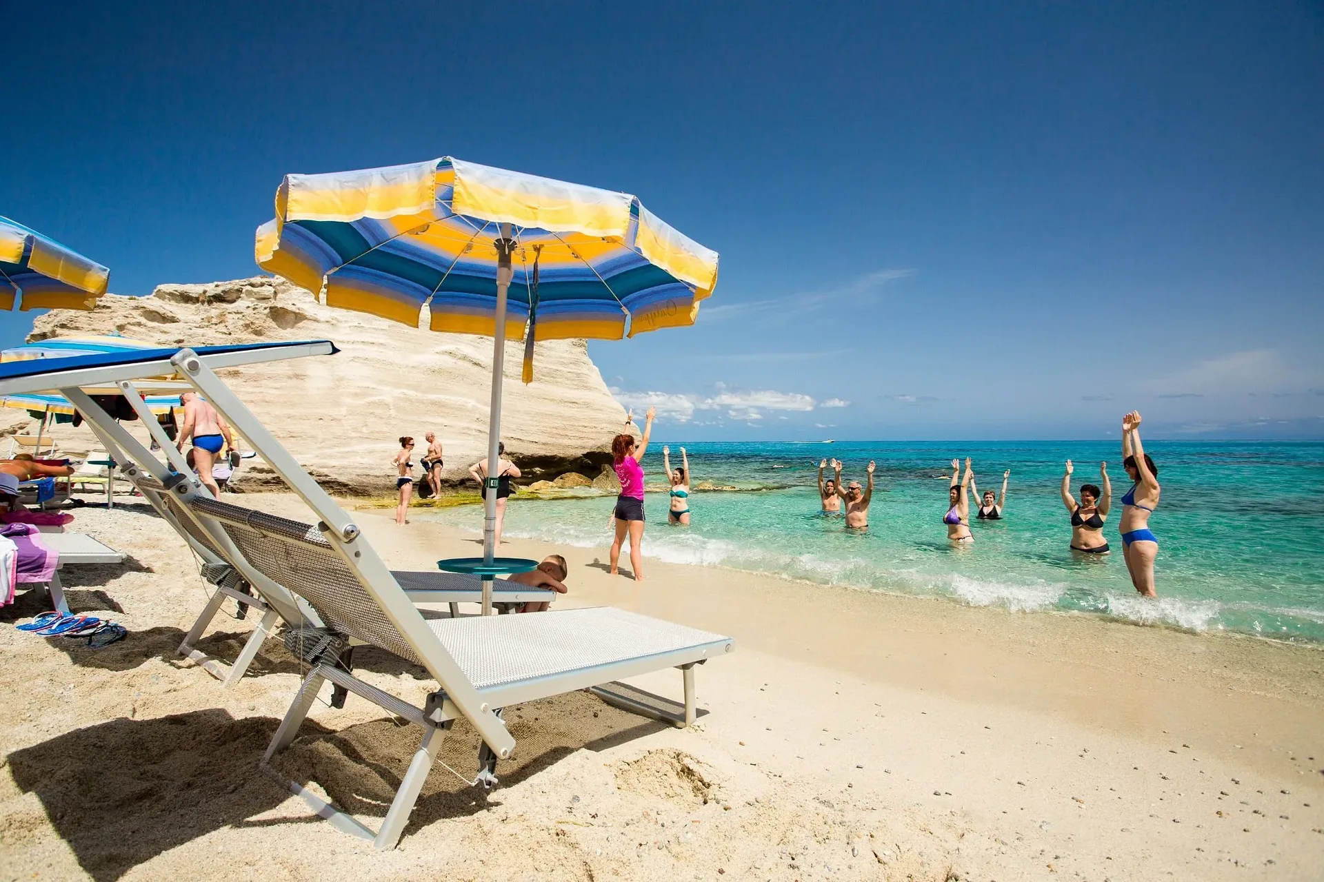 Bagnanti fanno ginnastica in acqua su spiaggia con ombrelloni e scogliera bianca