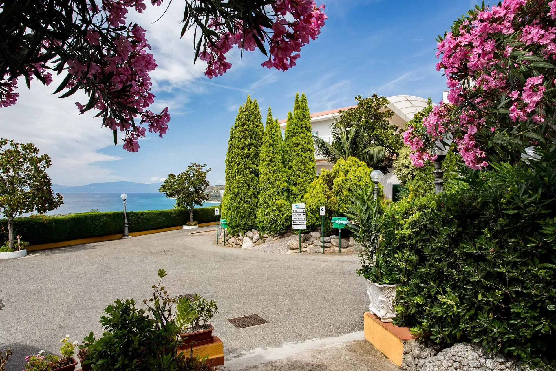 Vialetto d'ingresso dell'hotel con fiori rosa, cipressi e vista sul mare
