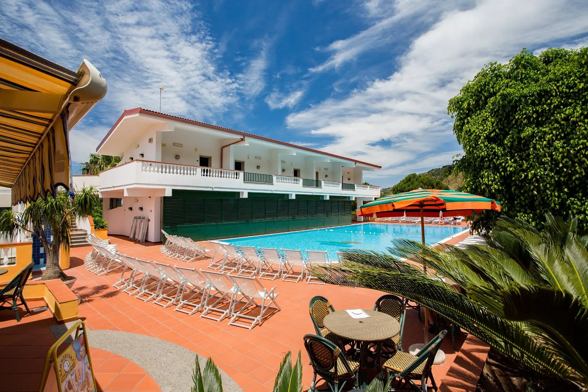 Esterno dell'hotel con piscina, lettini e palme sotto il cielo azzurro