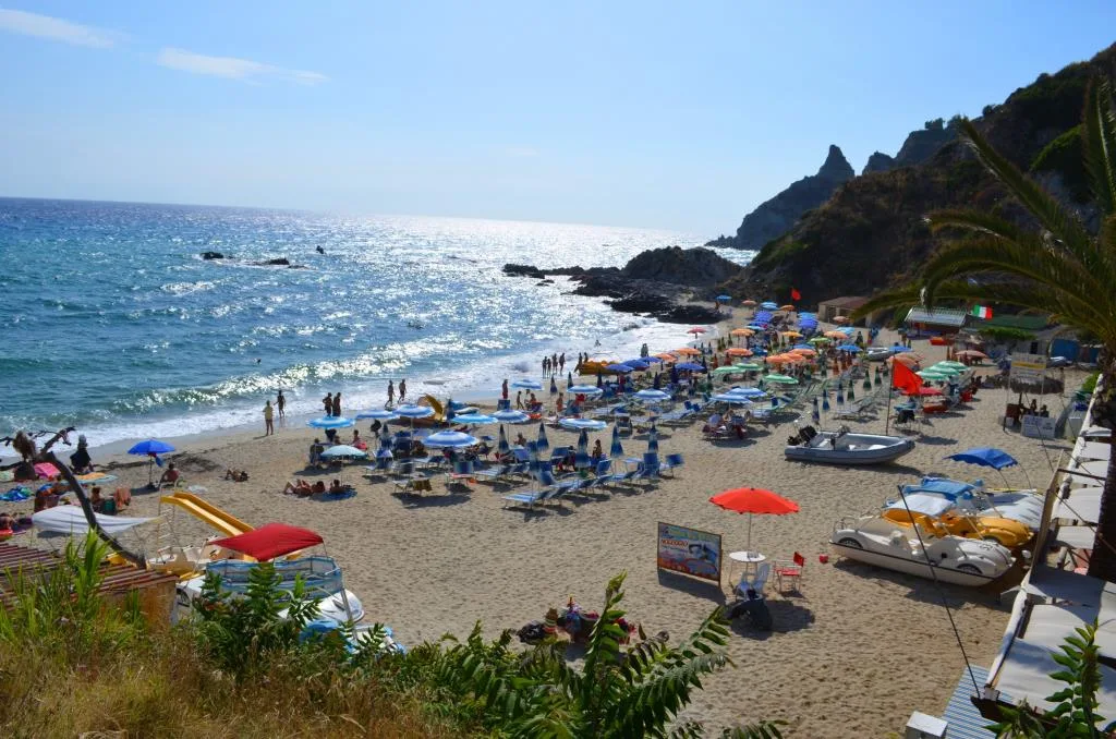 Spiaggia affollata con ombrelloni colorati, mare cristallino e scogliera