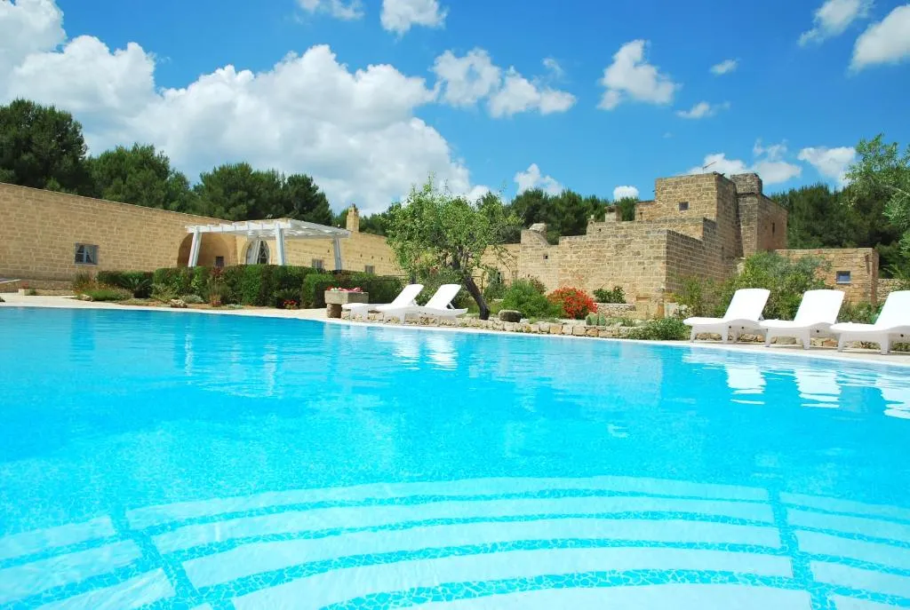 Ampia piscina azzurra con lettini bianchi e masseria storica circondata da pini