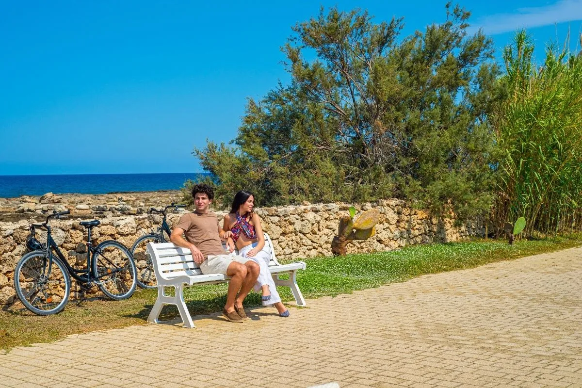 Coppia seduta su panchina lungo il percorso costiero con biciclette e vista mare