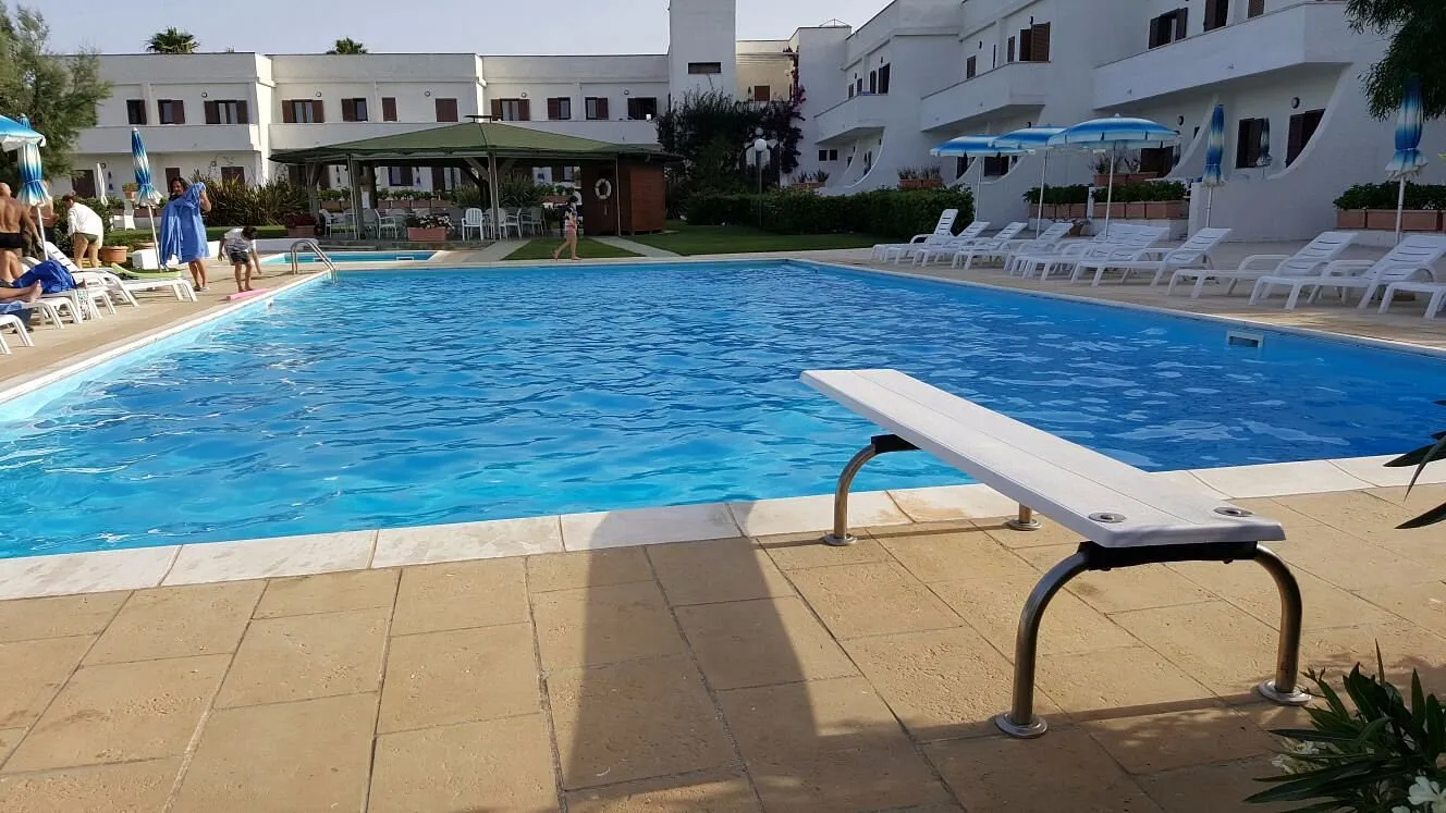 Piscina con trampolino in primo piano e lettini bianchi tra gli edifici bianchi del residence