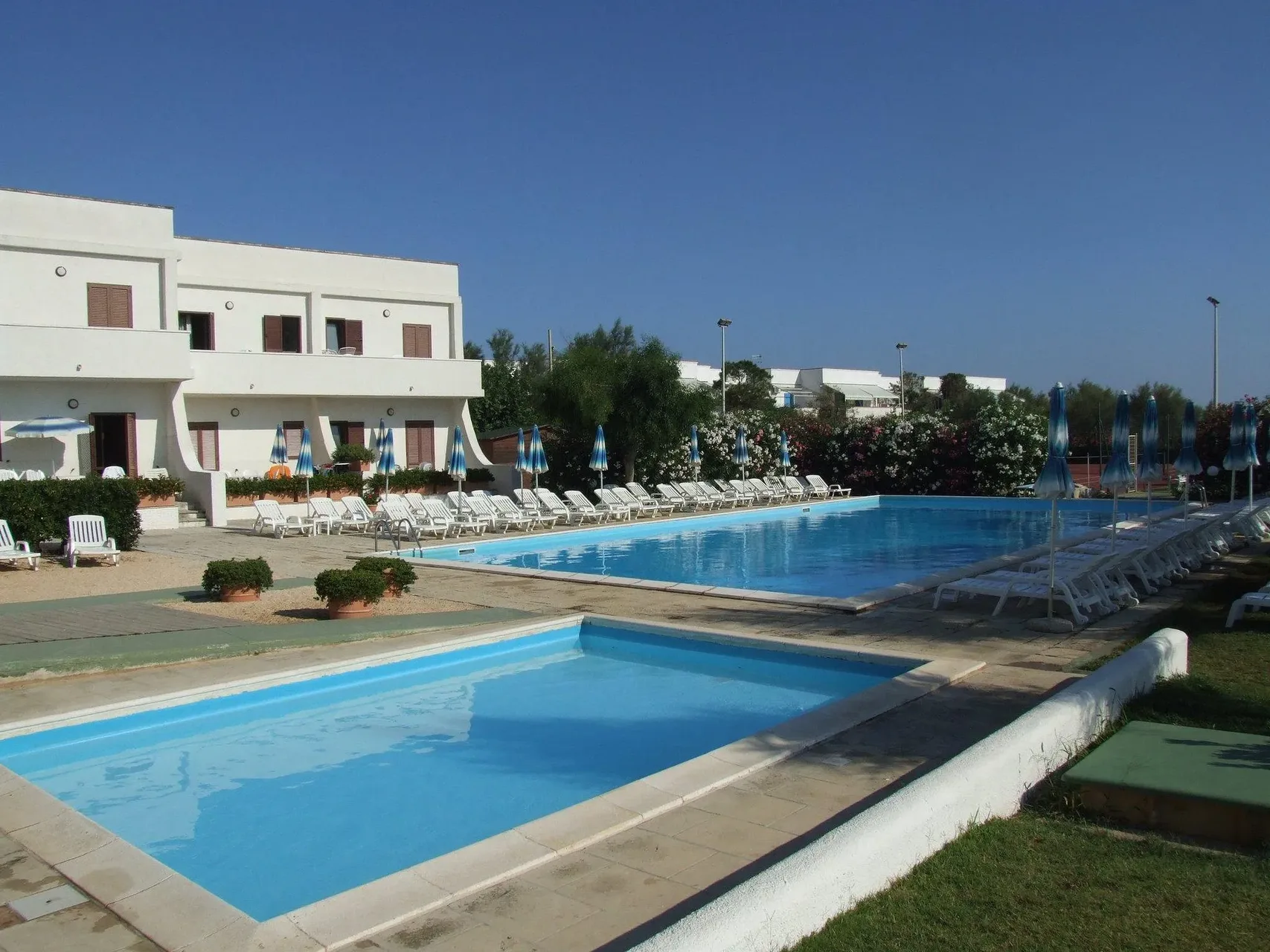 Due piscine di diverse dimensioni con lettini bianchi e ombrelloni accanto all'edificio bianco del residence