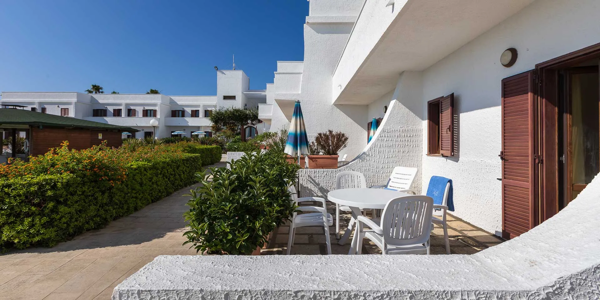 Terrazza esterna di un appartamento con tavolo e sedie bianche tra siepi e edifici bianchi