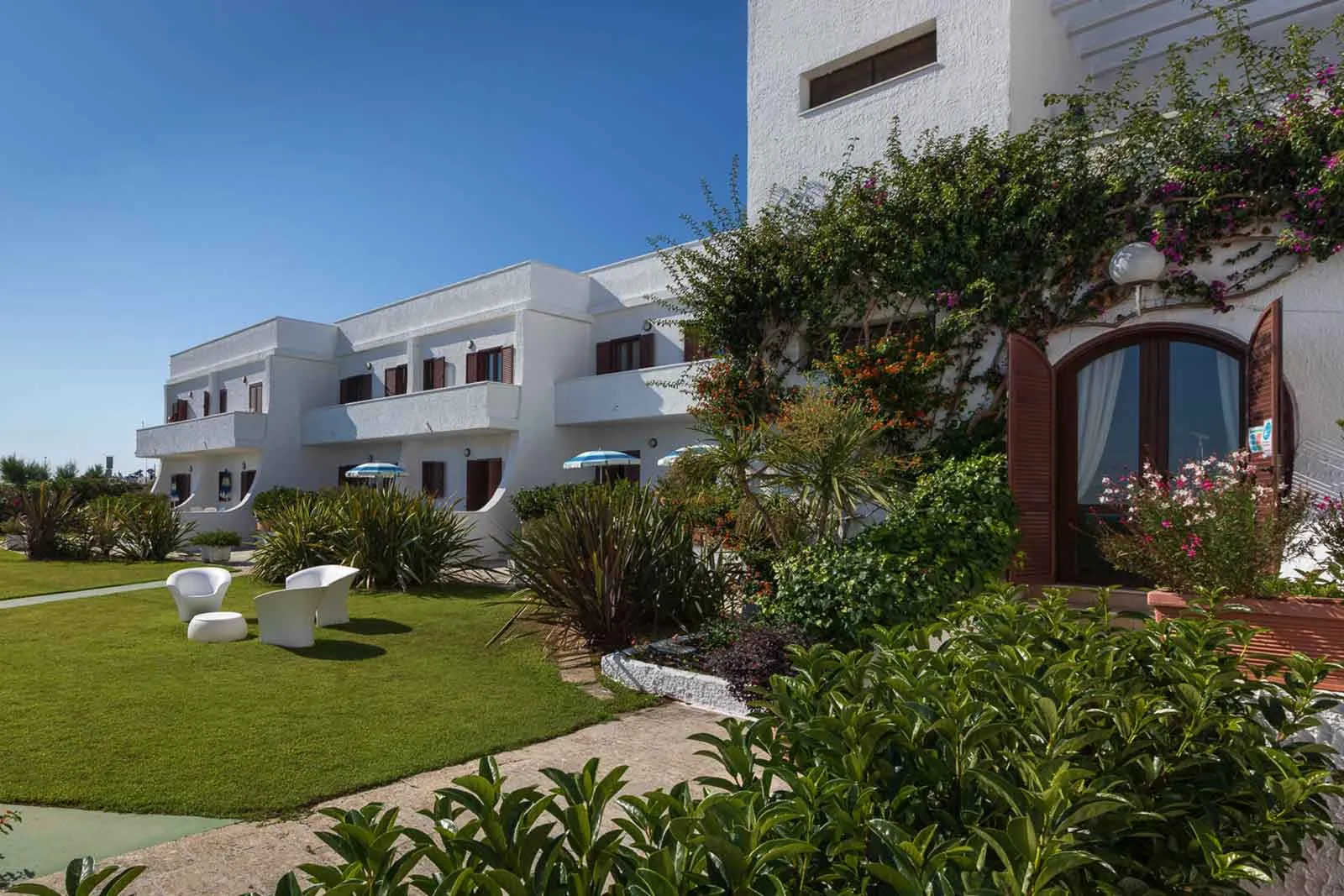 Edificio bianco del residence con giardino fiorito, bouganville e poltrone bianche sul prato
