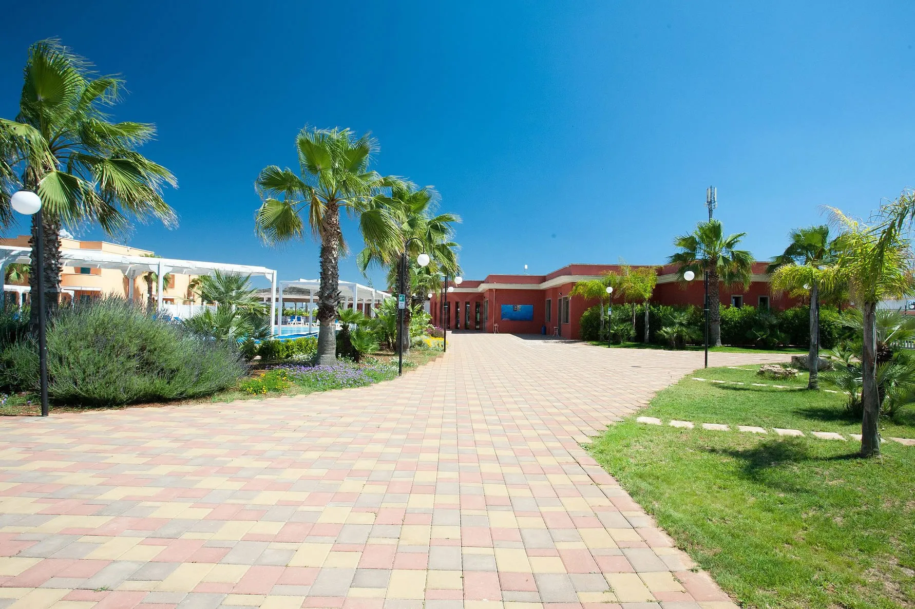 Vialetto pavimentato del resort tra palme e aiuole fiorite, con edificio rosso e piscina sullo sfondo