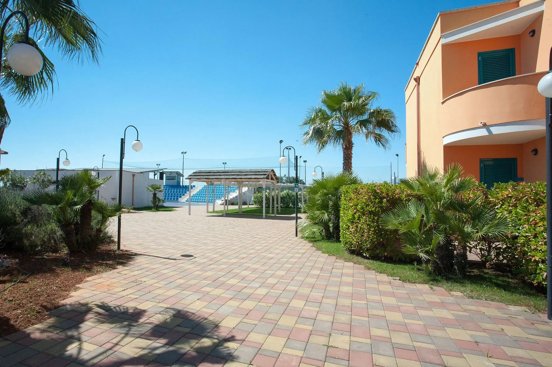Area esterna del resort con vialetto, palme e gradinate dell'anfiteatro, edificio color arancio a destra