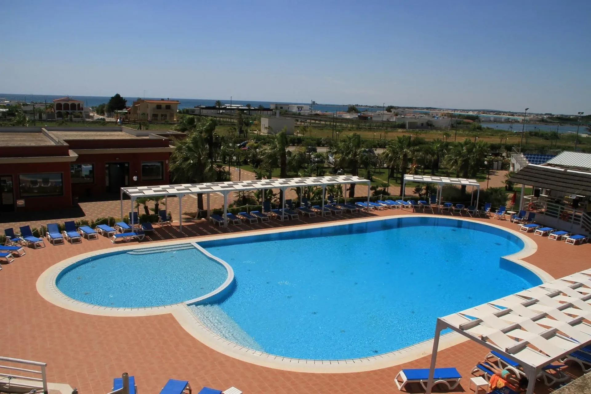Vista panoramica dall'alto della piscina con lettini blu e pergolati, paesaggio e mare sullo sfondo