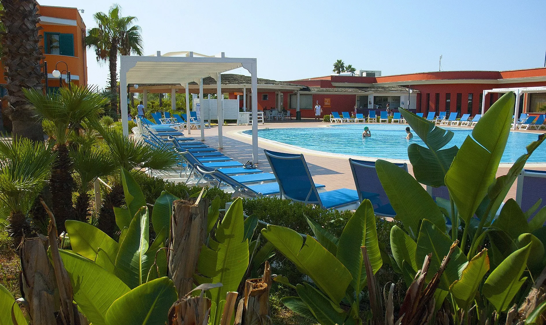 Piscina del resort vista tra piante tropicali, lettini blu e pergolati bianchi con edificio rosso sullo sfondo
