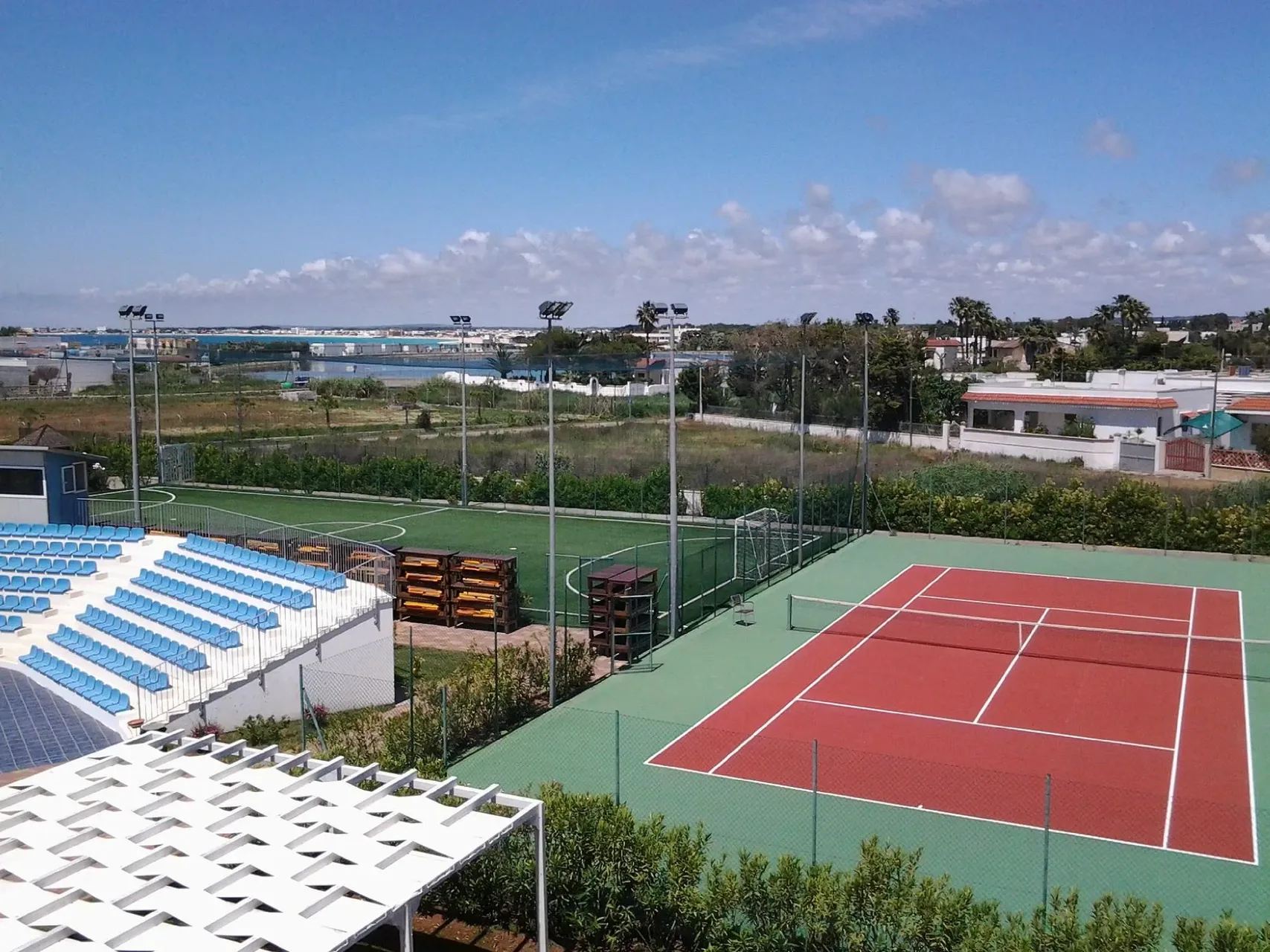 Vista dall'alto di campo da tennis e campo da calcio con gradinate azzurre, paesaggio e mare sullo sfondo