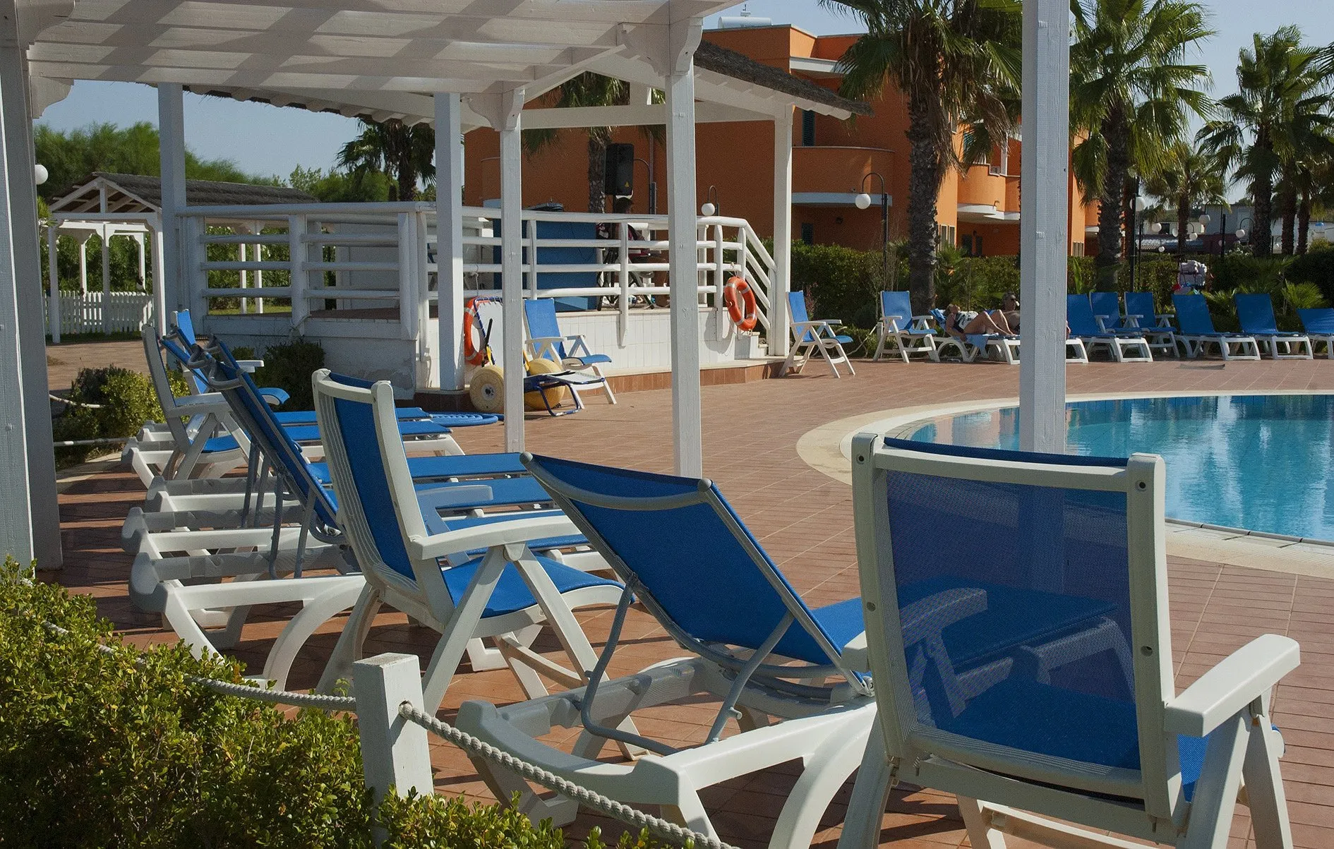 Lettini bianchi e blu a bordo piscina sotto pergolato, residence color arancio sullo sfondo