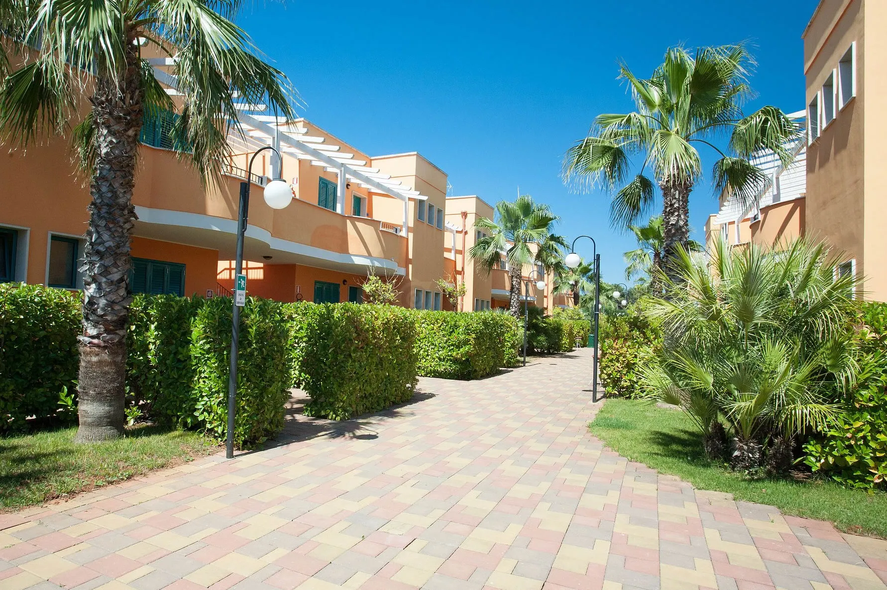 Vialetto pavimentato tra i residence color ocra con siepi e palme nel giardino del resort