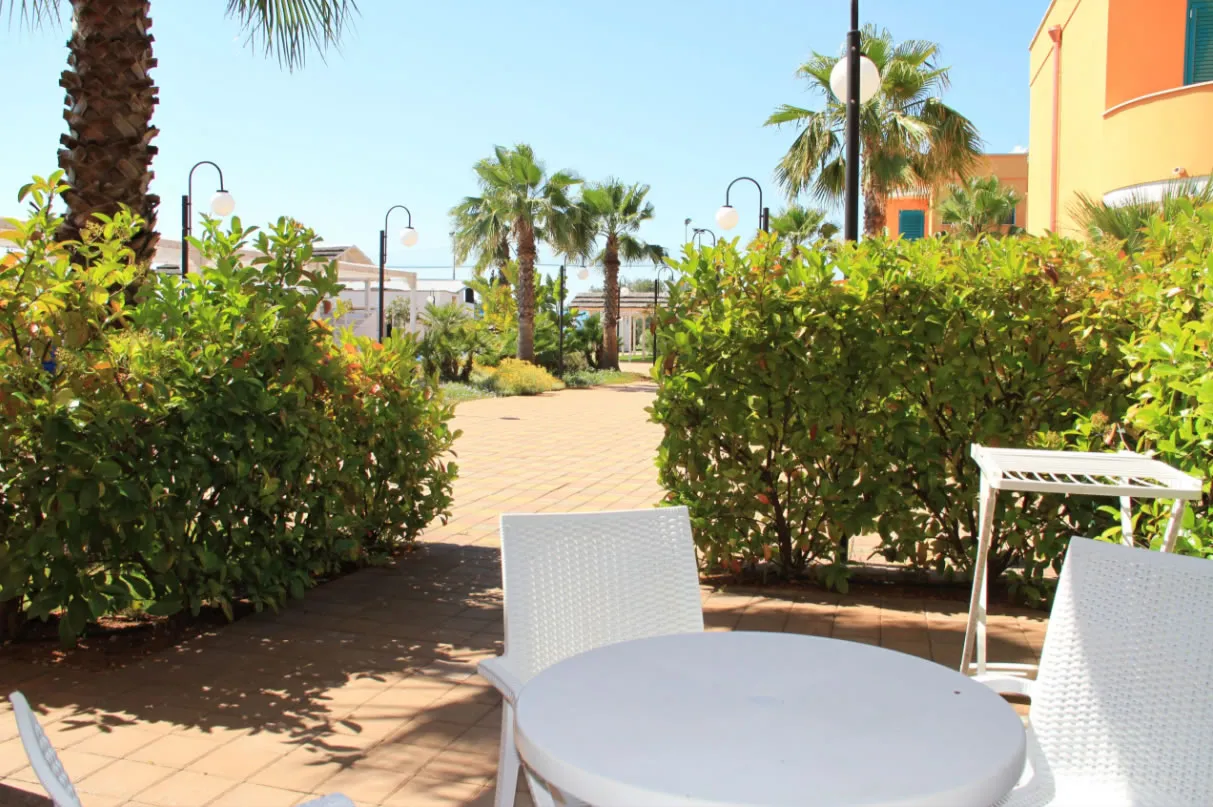 Patio esterno della camera con tavolo e sedie bianche affacciato su vialetto e palme