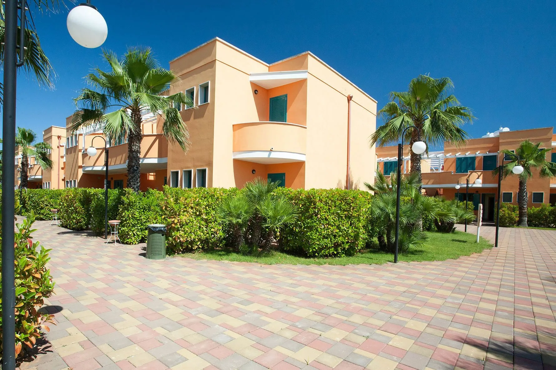 Esterno dei residence color ocra con balconi, siepi e palme sotto cielo azzurro