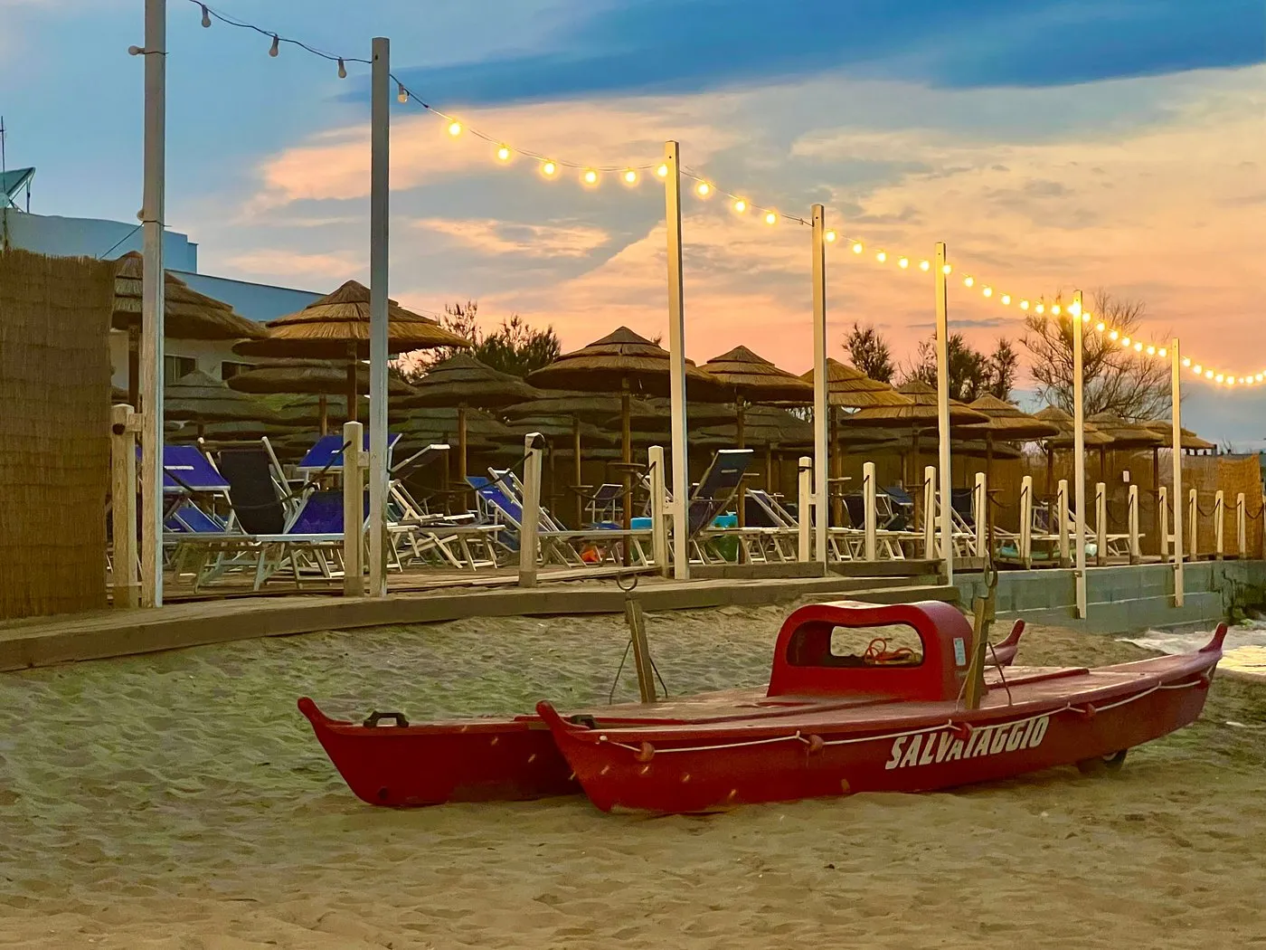 Spiaggia al tramonto con barca di salvataggio rossa e ombrelloni in paglia illuminati