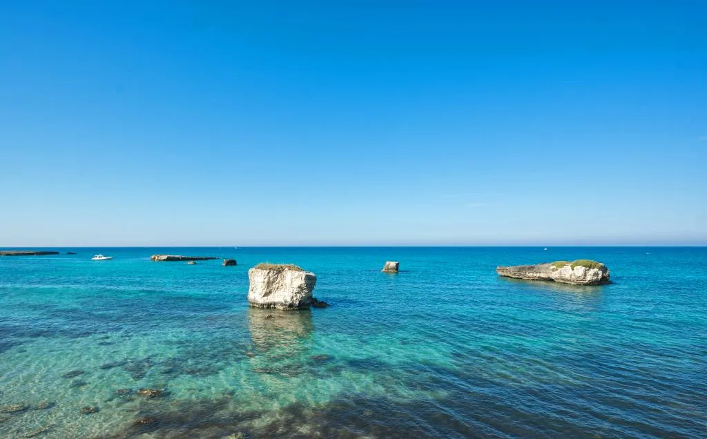 Mare azzurro limpido con faraglioni e piccole imbarcazioni sotto cielo sereno