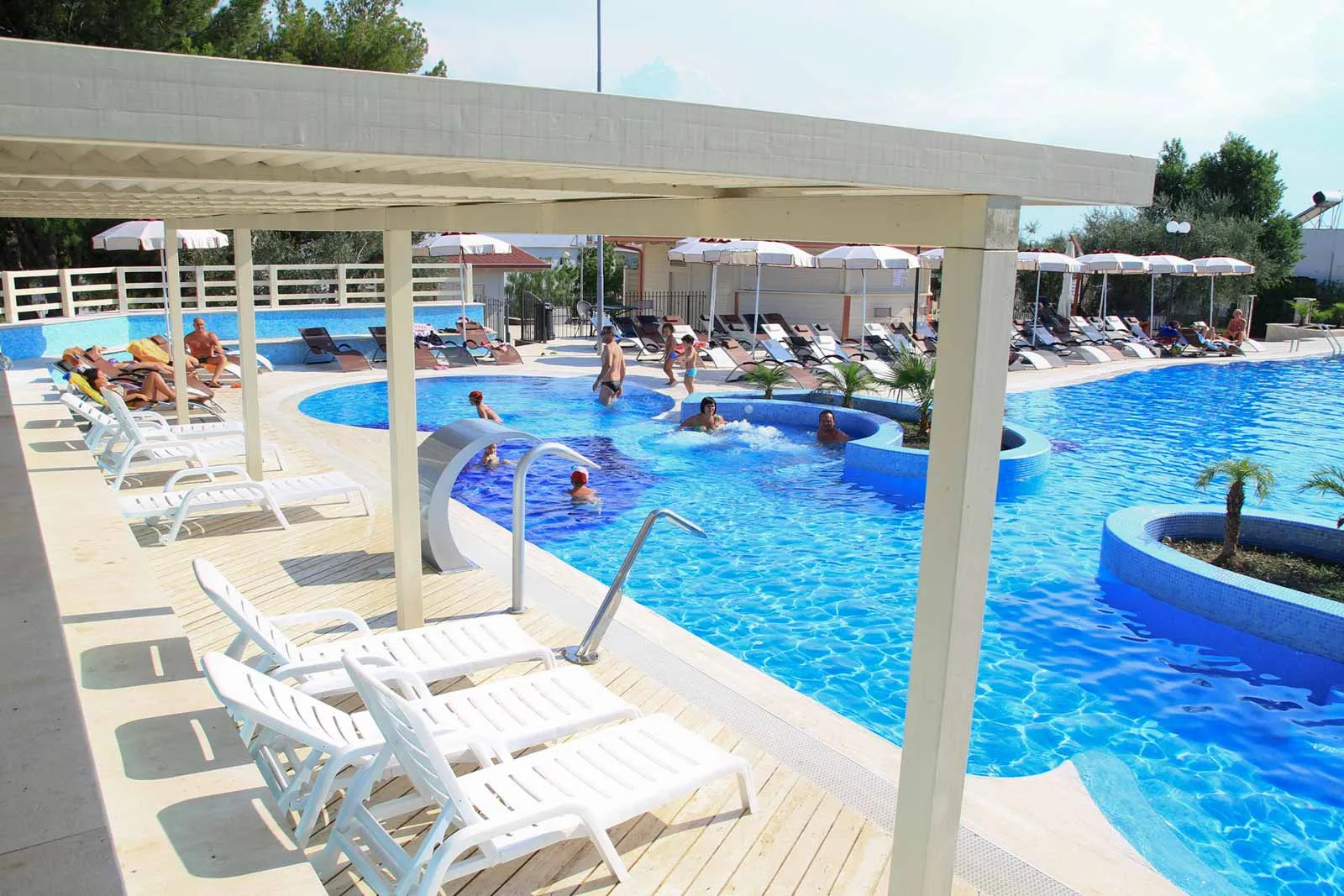Bordo piscina con lettini bianchi sotto pergolato e ospiti che fanno il bagno