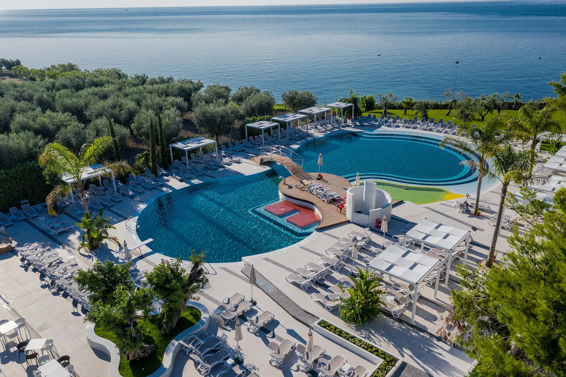 Piscina sinuosa con ponte in legno e lettini affacciata sul mare blu