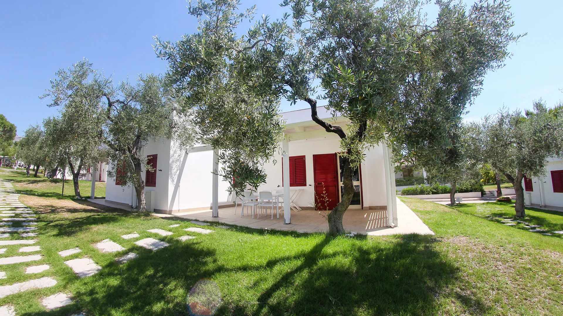 Bungalow bianco con persiane rosse e veranda con tavolo immerso tra gli ulivi